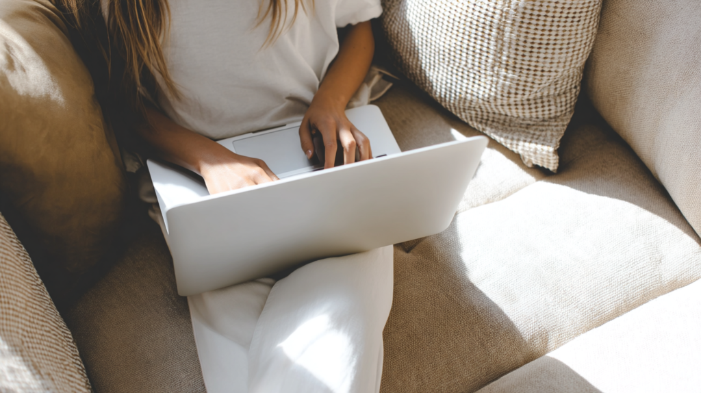 woman-working-couch a woman sits on a cozy beige sofa in the sunlight typing on a laptop