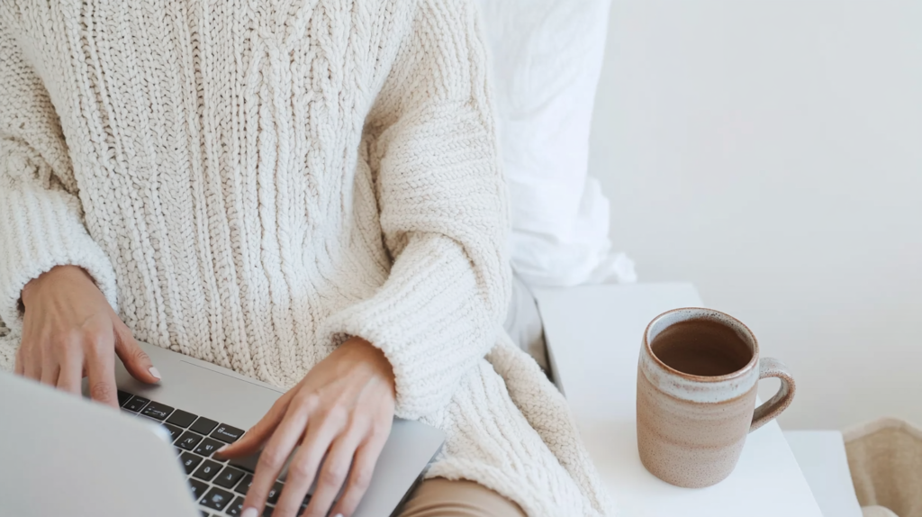 a woman in a white sweater works at a laptop with a mug of tea