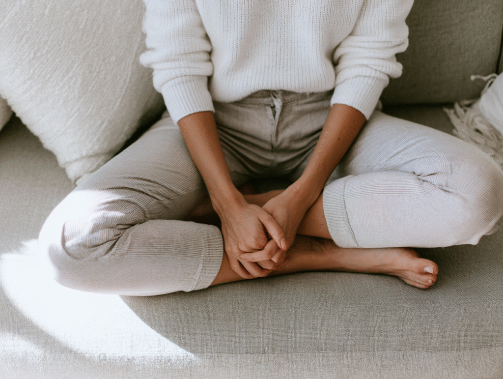 a woman sits cross legged on a couch in a sweatshirt and jeans
