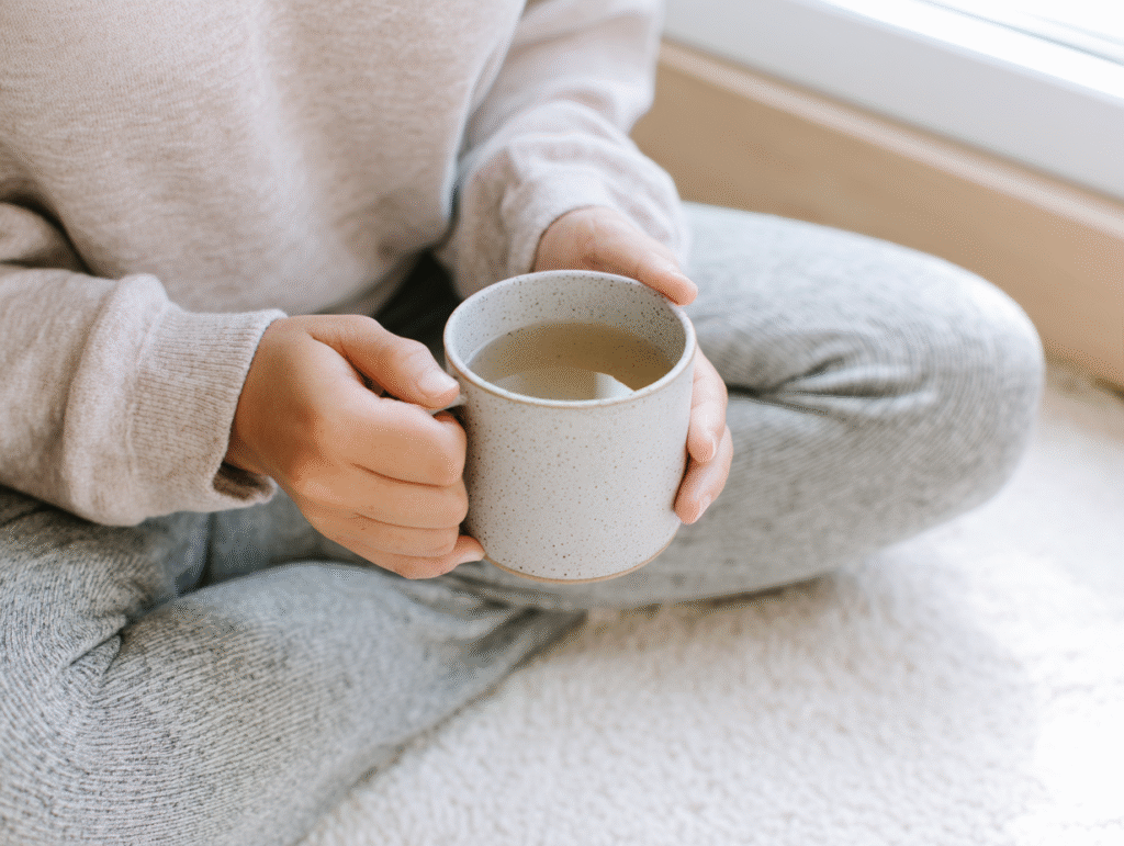 a woman sits cross legged holding a mug of tea with both hands