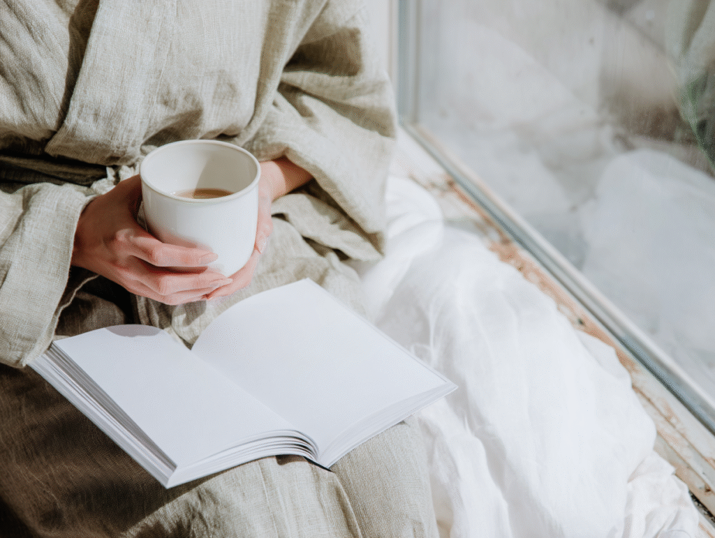 a woman holds a teacup and journal next to a window