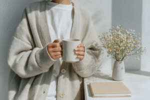 Person holding ceramic mug in cozy cardigan with wildflowers and notebook on table.