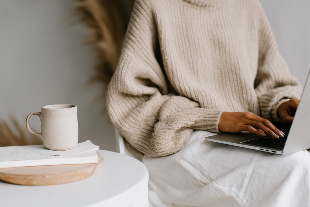 Person typing on a laptop wearing a beige sweater, with a coffee mug on a table nearby. Cozy workspace setting.