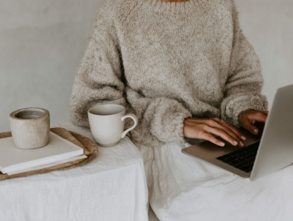 how-to-use-pinterest-laptop5 Cozy workspace with person in sweater typing on laptop, next to coffee mugs and books on a textured surface.