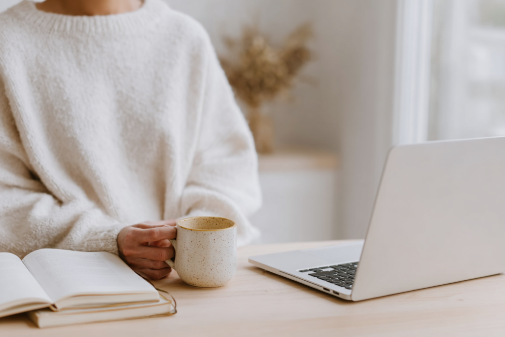 how-to-use-pinterest-mug Woman in cozy sweater holding mug, sitting at desk with laptop and book.