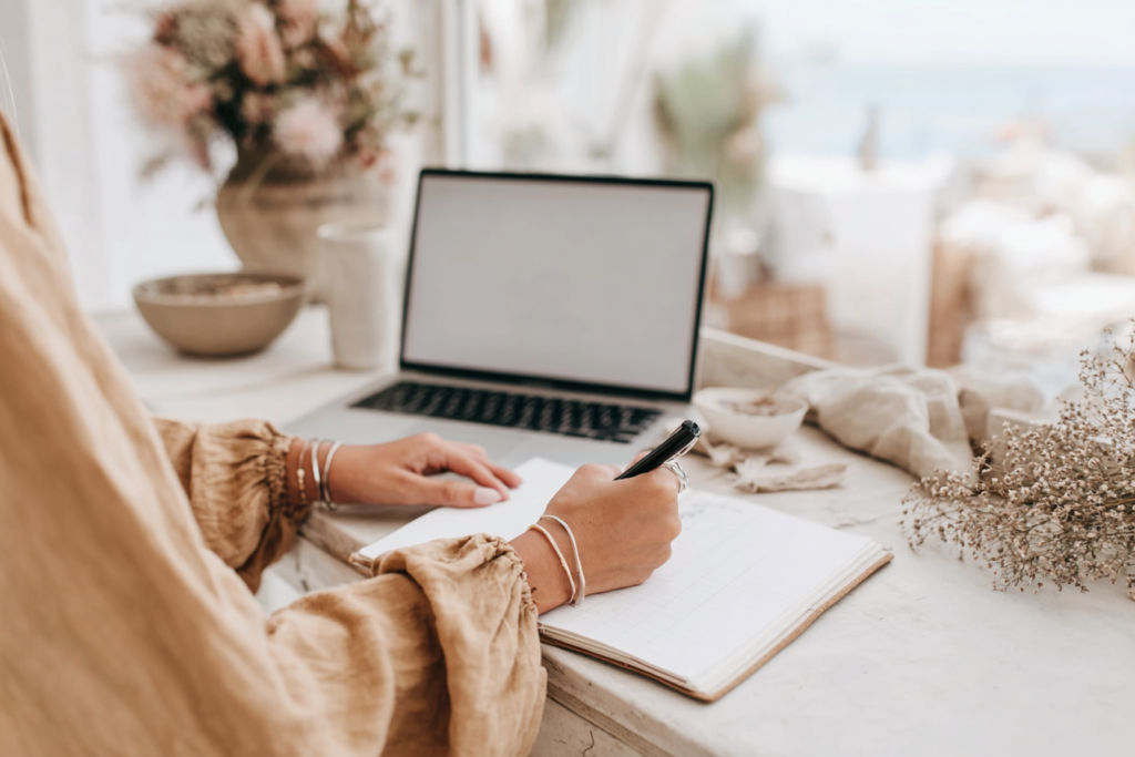 how-to-use-pinterest-notes Person writing in notebook at rustic desk; open laptop and cozy decor in background.