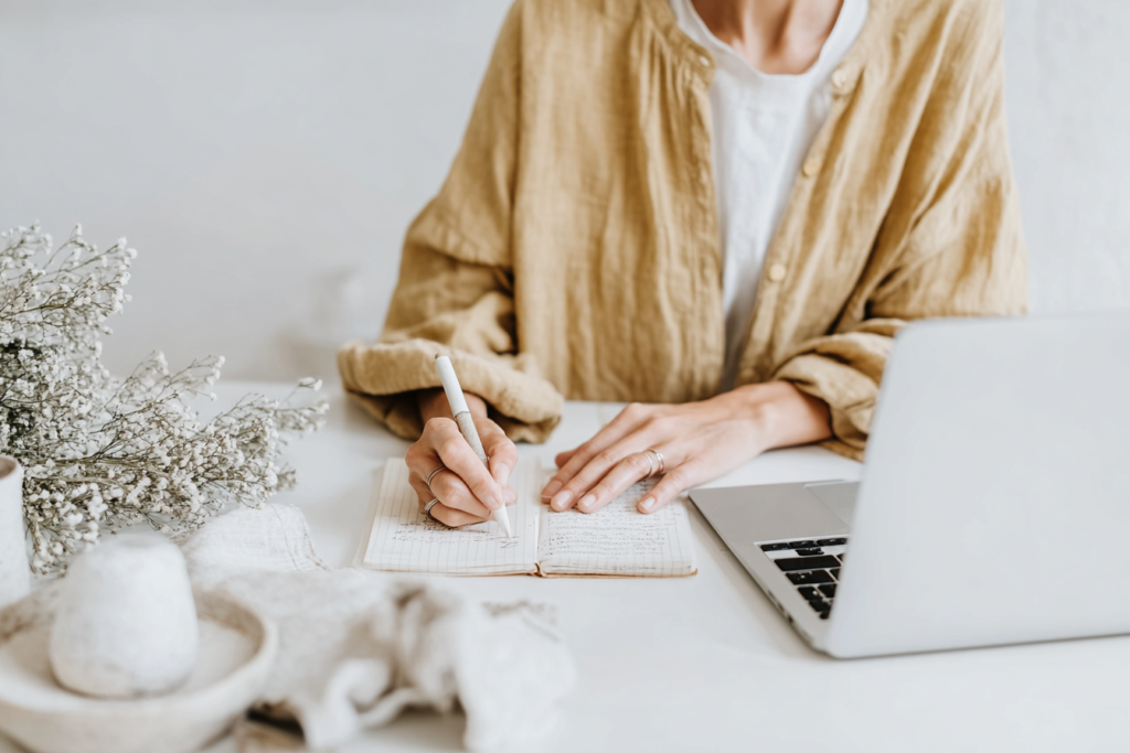 how-to-use-pinterest-notes3 Person writing in a notebook beside a laptop on a white desk, with decorative flowers nearby.