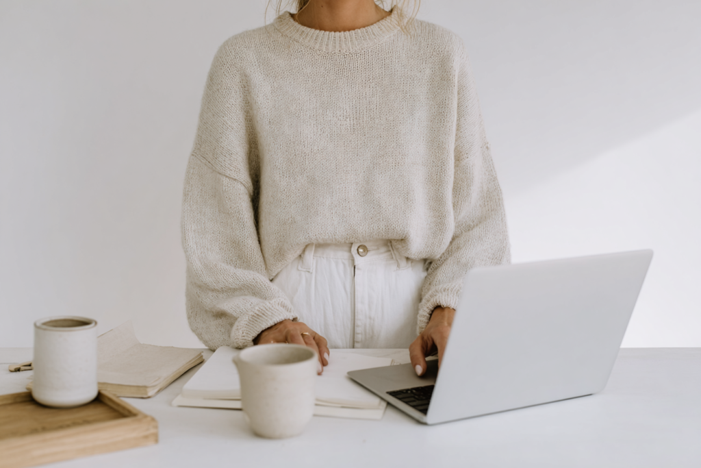 Person in a cozy sweater working on a laptop with coffee cups and notebooks on the desk. Minimalist workspace.