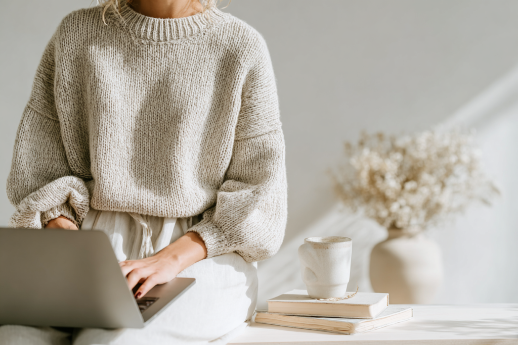 Woman in cozy sweater using laptop, with coffee mug and books on table, in sunlight.