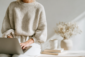 Woman in cozy sweater using laptop, with coffee mug and books on table, in sunlight.
