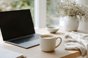 Cozy workspace with laptop, coffee mug, and soft blanket on wooden desk by window.