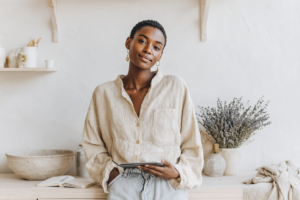 Person in a casual outfit holding a tablet in a cozy, minimalist room with a shelf and plants in the background.