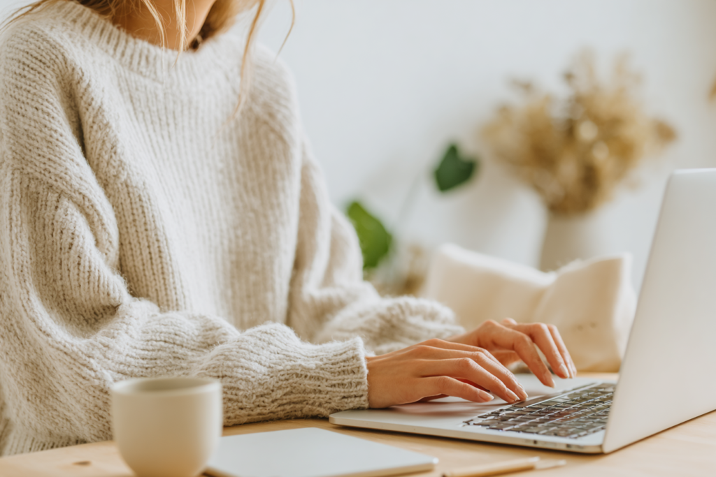 DBS-working-2-26-5 Person typing on a laptop at a cozy workspace with a coffee mug, wearing a soft sweater.