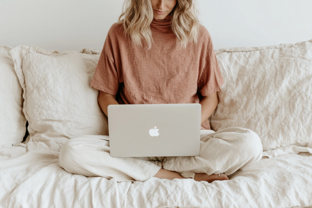 Person working on a laptop while sitting on a cozy bed with neutral bedding. Relaxed home workspace atmosphere.