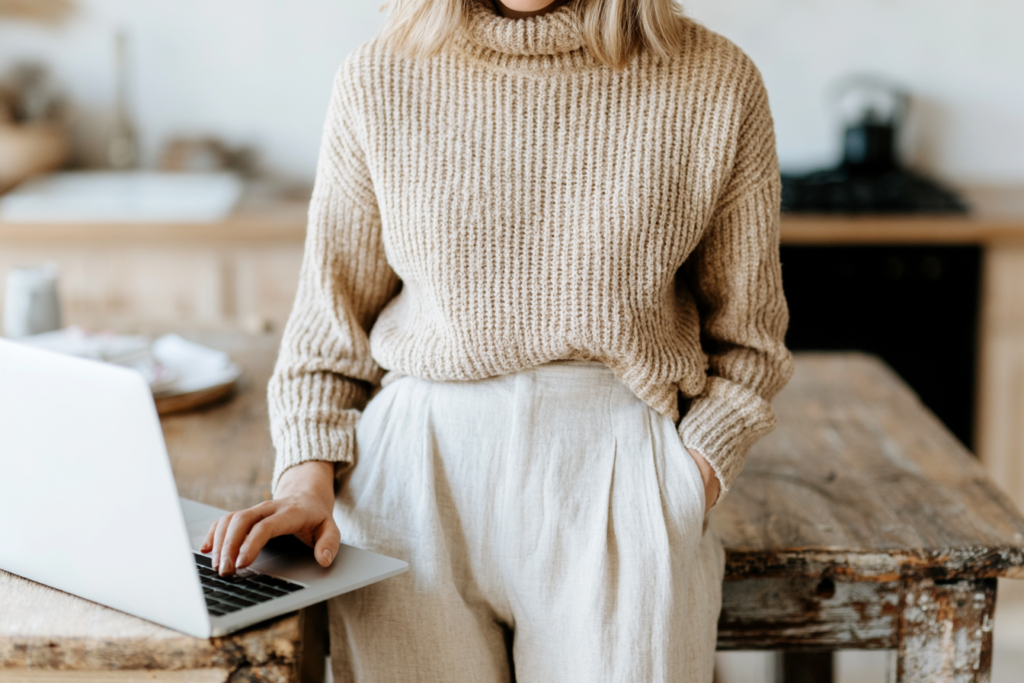 Person in a cozy beige sweater using a laptop in a rustic kitchen setting.