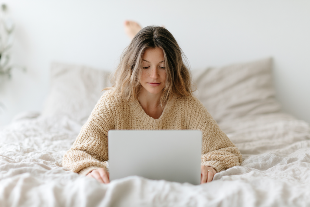 Woman in cozy sweater using a laptop on bed, focused on screen, light background.