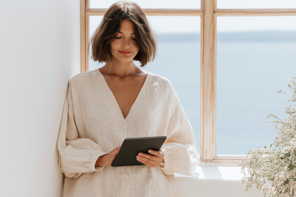Woman reading a tablet by a sunny window, wearing a light beige blouse.