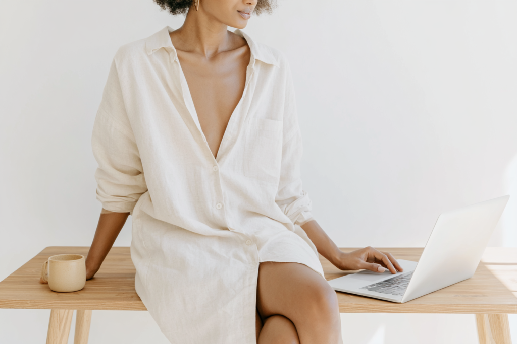 Person in beige shirt sitting on wooden desk, working on a laptop with a mug nearby. Minimalist home office setting.