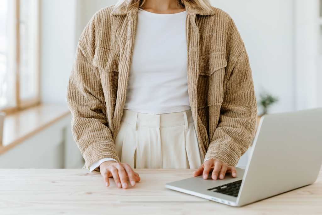 Person in beige outfit standing at a desk with an open laptop, ready for work or study in a bright room.