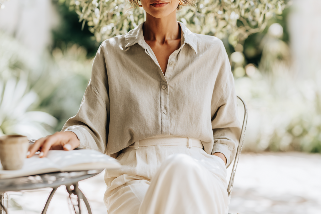 Woman in beige outfit sitting outside, holding a book and coffee, with greenery in the background.
