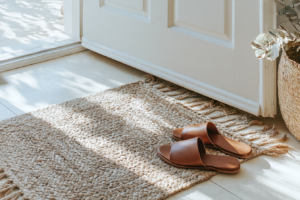 Brown leather sandals on a woven jute rug by a sunlit door, accompanied by a wicker basket and eucalyptus.
