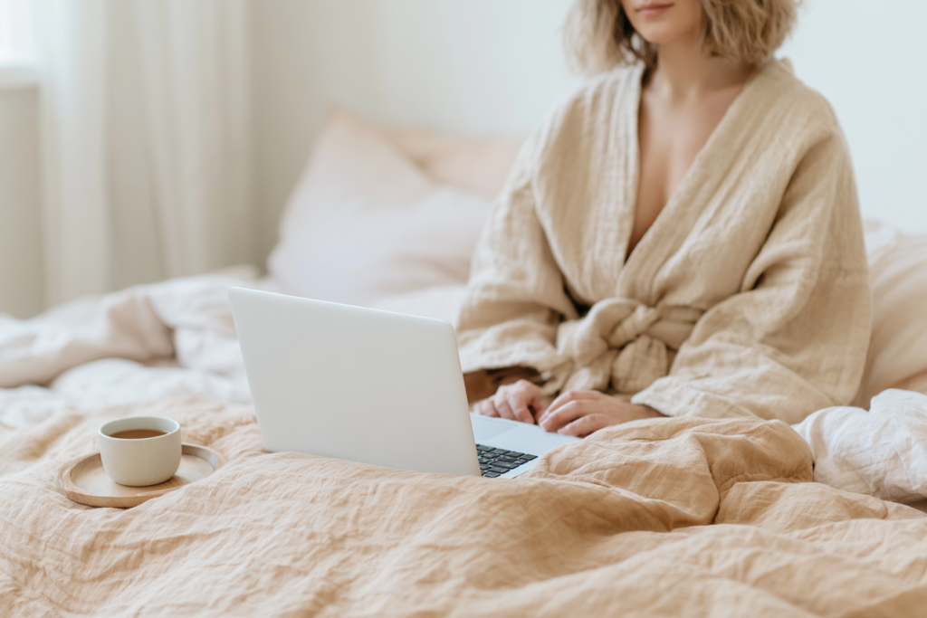 Person in cozy robe working on laptop in bed with coffee, minimalist bedroom, work-from-home morning routine.