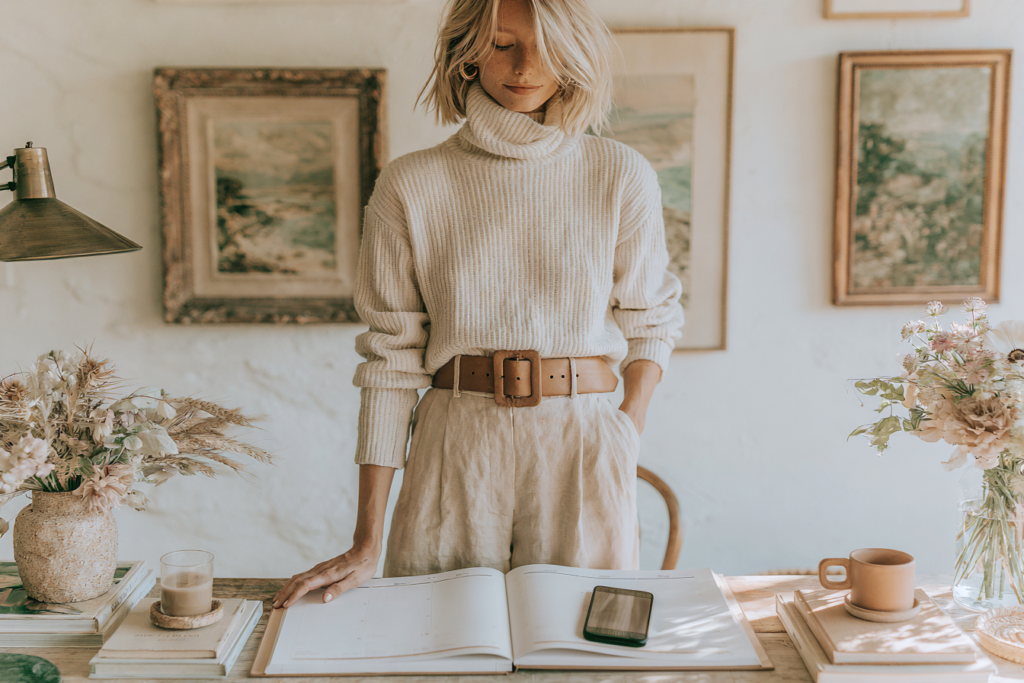 desk-book-gallery-wall-2 Minimalist home office scene: person in beige sweater plans schedule at wooden desk with flowers, coffee, and smartphone.