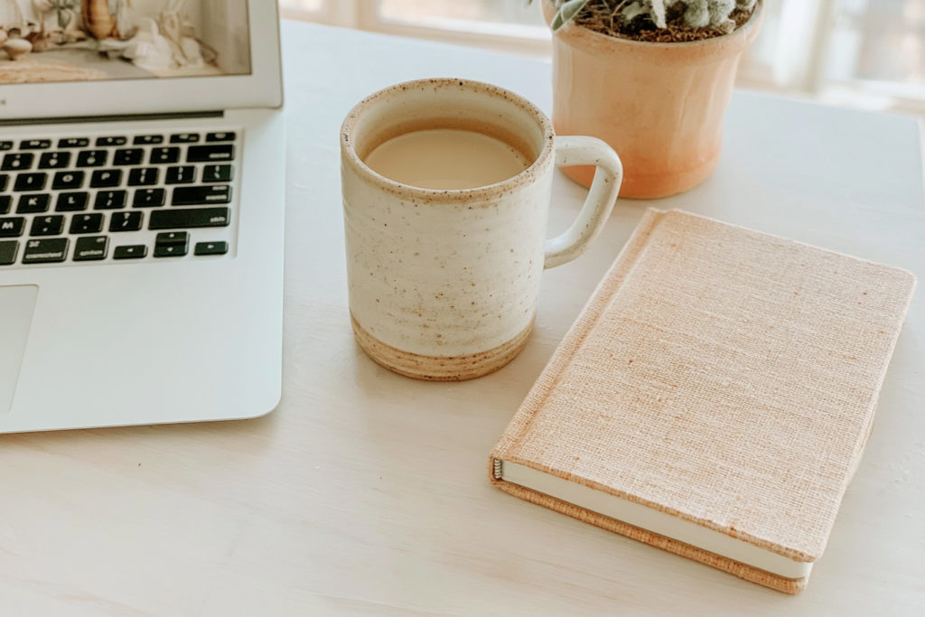 Cozy home office desk with coffee in ceramic mug, laptop, linen journal notebook, and potted plant in natural light