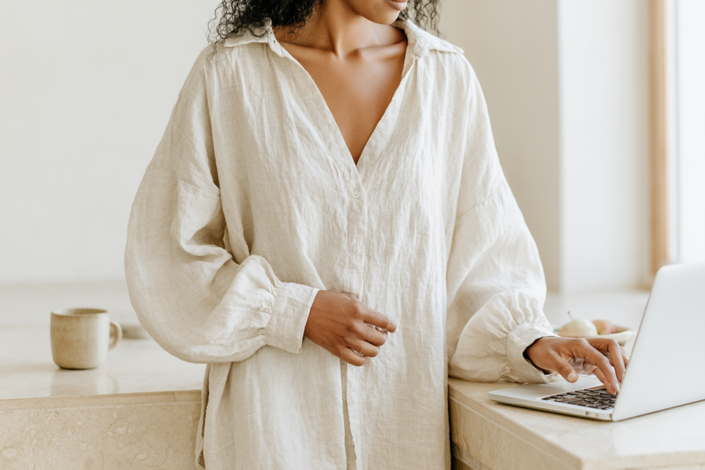 Person in beige linen shirt working on laptop in bright minimalist kitchen, coffee mug nearby — remote work home office.