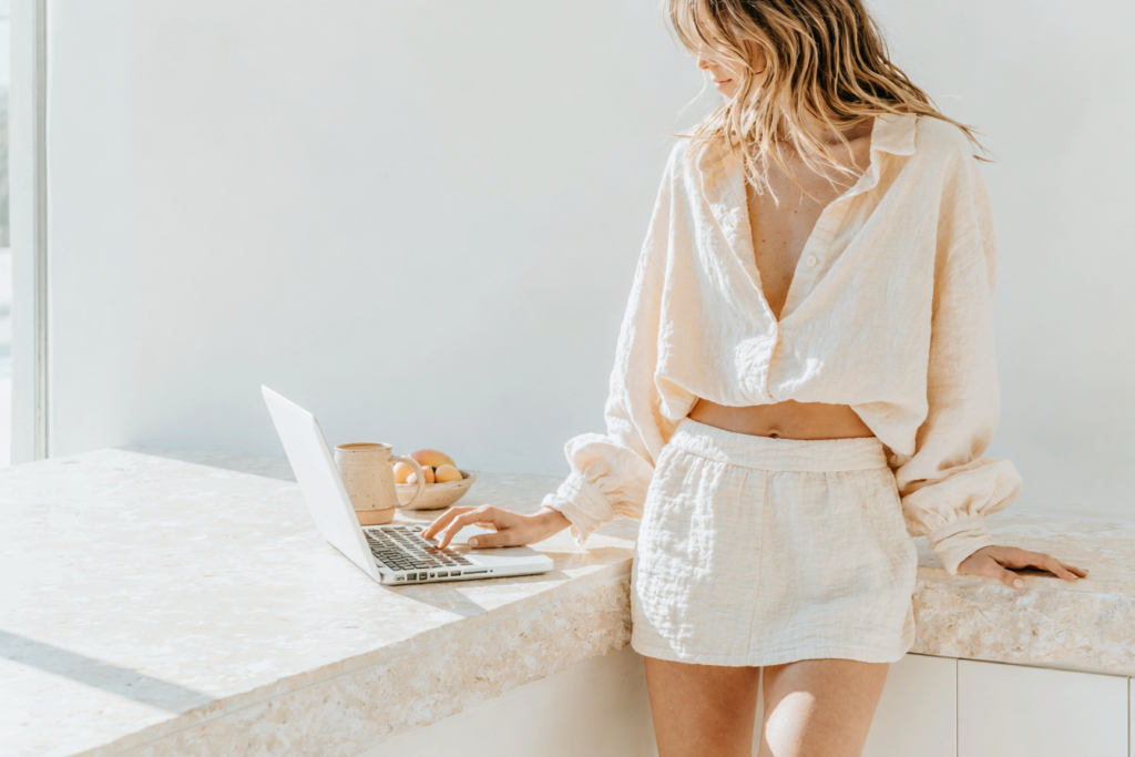 Woman in linen loungewear working on laptop in sunlit minimalist kitchen, modern home office vibe