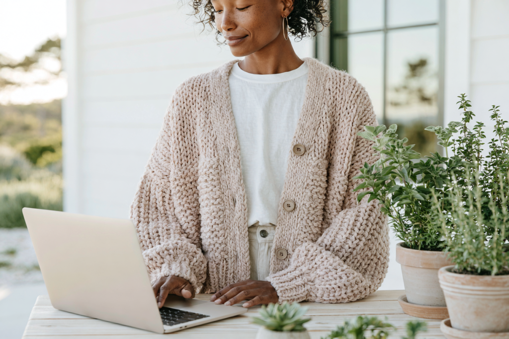 Person in beige chunky knit cardigan working on laptop at patio workspace with potted herbs and succulents.
