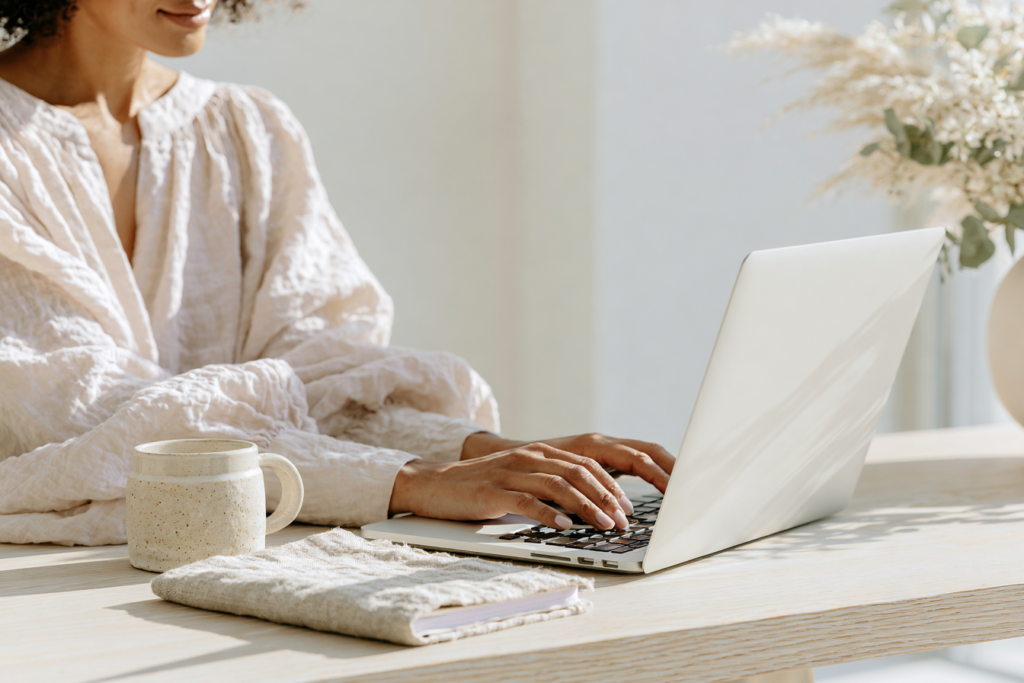 Person typing on laptop at sunlit home office desk with coffee mug and notebook, minimalist remote work setup.