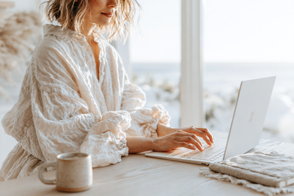 Person typing on laptop in bright home office, linen shirt, coffee mug and notebook on wooden desk, cozy remote work scene.