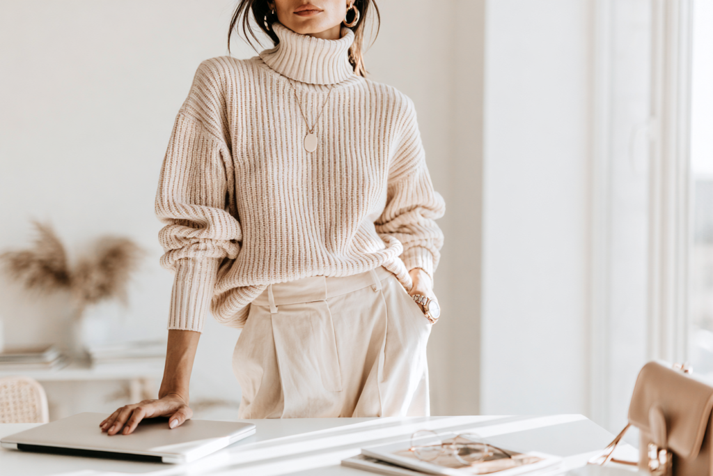 Woman in ribbed knit turtleneck sweater and high-waist trousers standing by bright desk with laptop in neutral office