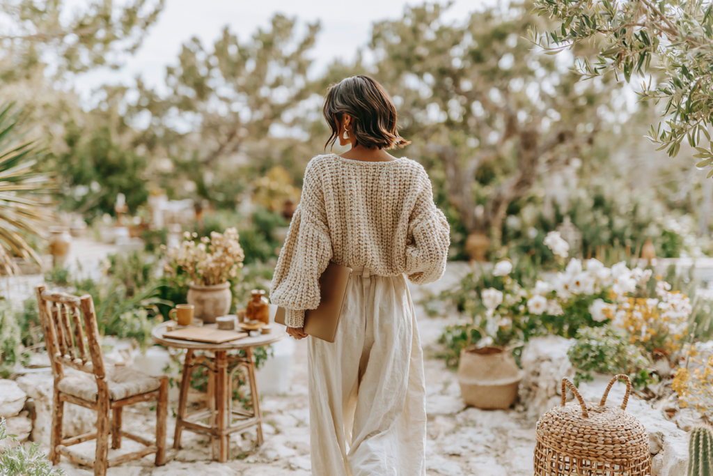 Woman in chunky knit sweater holding laptop walks through rustic garden patio with flowers and wicker decor.